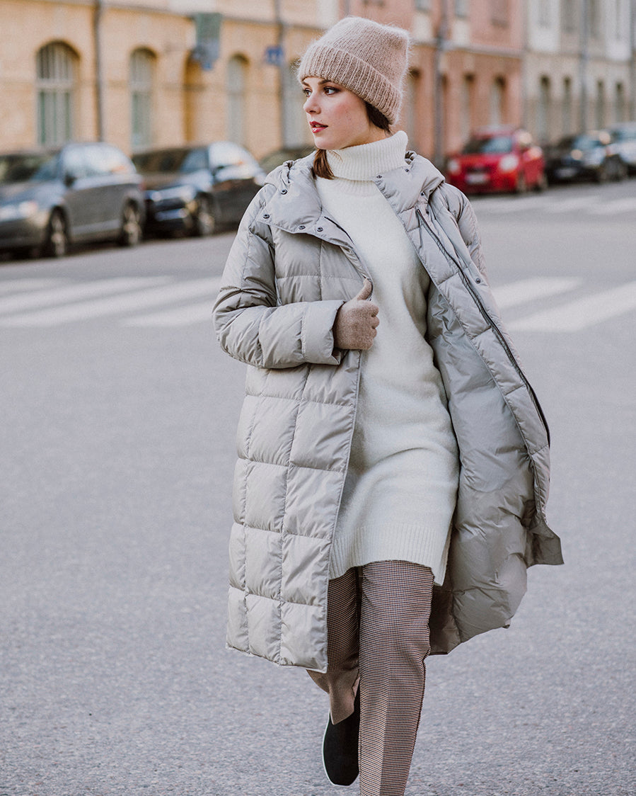 Nainen kävelee kadulla yllään auki oleva vaalea, pitkä Essi-kevytuntuvatakki (väri: samppanja), jossa on neliömäiset tikkaukset. Asuun kuuluu myös beige pipo, vaalea neulemekko, raidalliset housut ja beiget hanskat. Taustalla näkyy kaupunkimaisemaa ja pysäköityjä autoja.