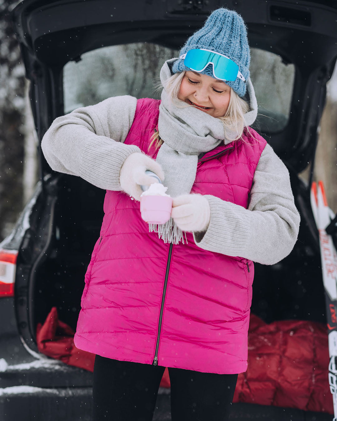 Nainen seisoo auton takakontin edessä ja kaataa juomaa mukiin. Hänellä on yllään pinkki Mesi-untuvaliivi (väri: pitaija), harmaa neule ja sininen pipo, sekä laskettelulasit otsalla.