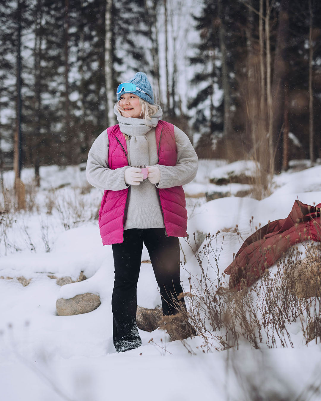 Nainen seisoo lumisessa metsämaisemassa yllään pinkki Mesi-untuvaliivi (väri: pitaija), harmaa neule ja mustat housut. Hänellä on sininen pipo ja aurinkolasit, ja hän katsoo hymyillen sivulle.