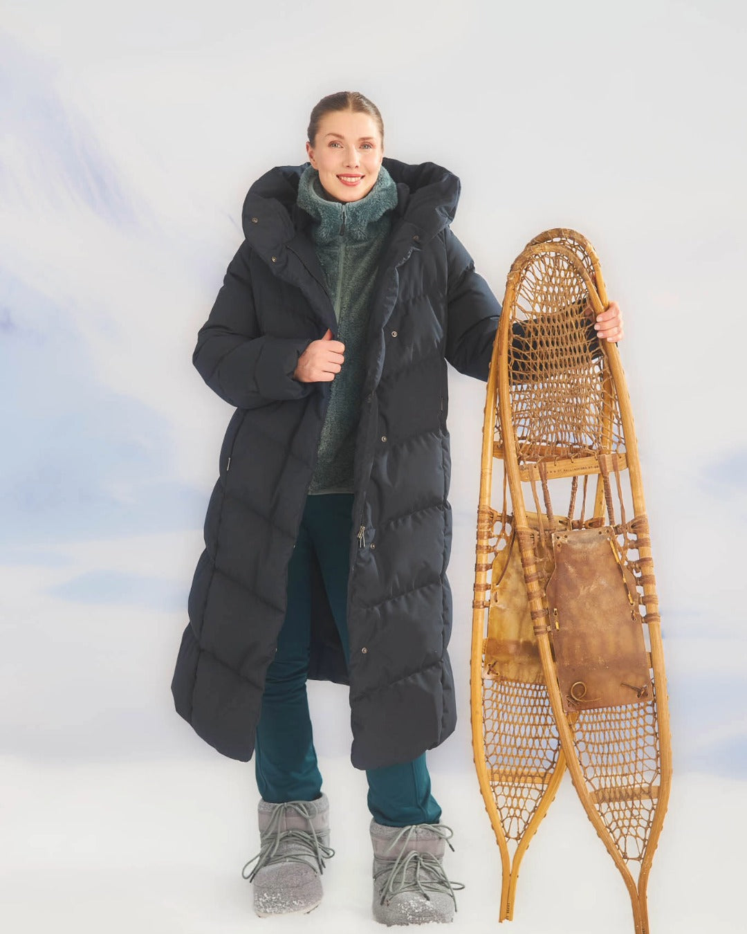 Naisella on yllään pitkä Aino untuvatakki mustana (väri: mustatorvisieni.) Hän pitää vieressään vanhanaikaisia lumikenkiä.