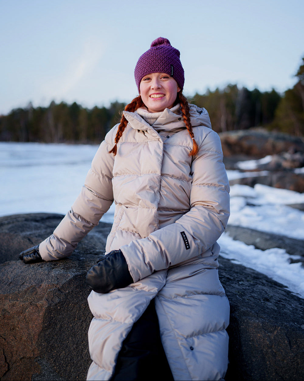 Nainen istuu suurella rantakivellä talvisessa maisemassa, taustalla jäinen järvi ja metsää. Hänellä on yllään vaalea Joutsenen Blanka-untuvatakki (väri: dyyni), violetti pipo ja mustat lapaset. Hän hymyilee ja katsoo kameraan.