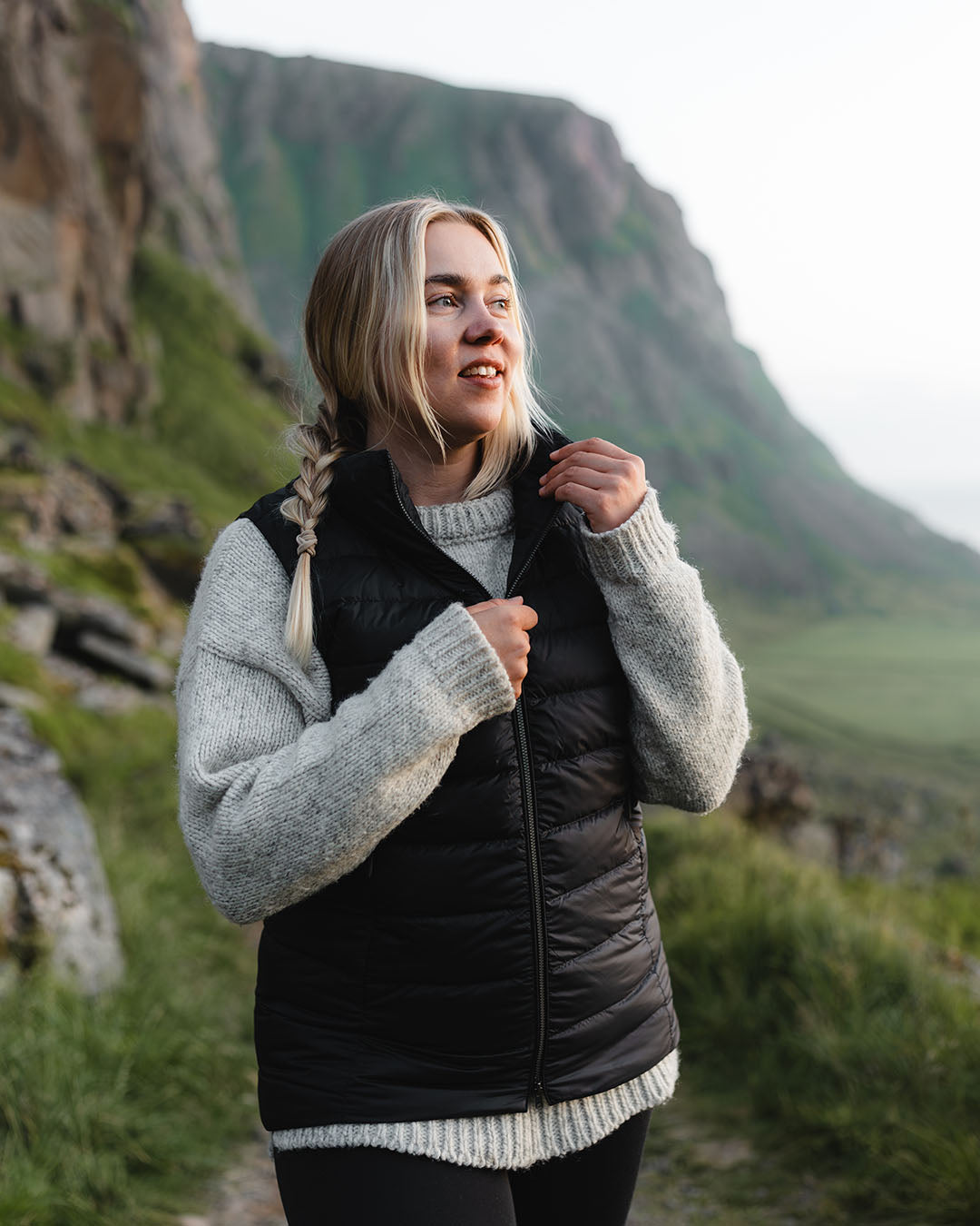 Nainen seisoo vuoristomaisemassa yllään musta Lady-untuvaliivi (väri: hiilenharmaa) ja harmaa villapaita. Hän katsoo sivulle ja pitää liivin vetoketjusta kiinni.