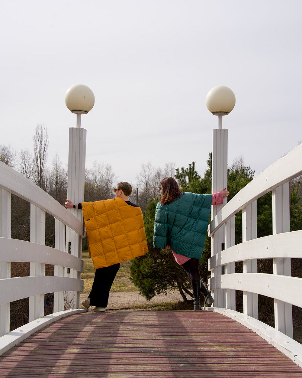 Kaksi henkilöä seisoo kaarisillalla selin kameraan, toinen pukeutuneena keltaiseen (väri: meripihka) ja toinen vihreään (väri: vihreä paprika) Otava-untuvaponchoon. Taustalla näkyy keväinen maisema, jossa on puita.