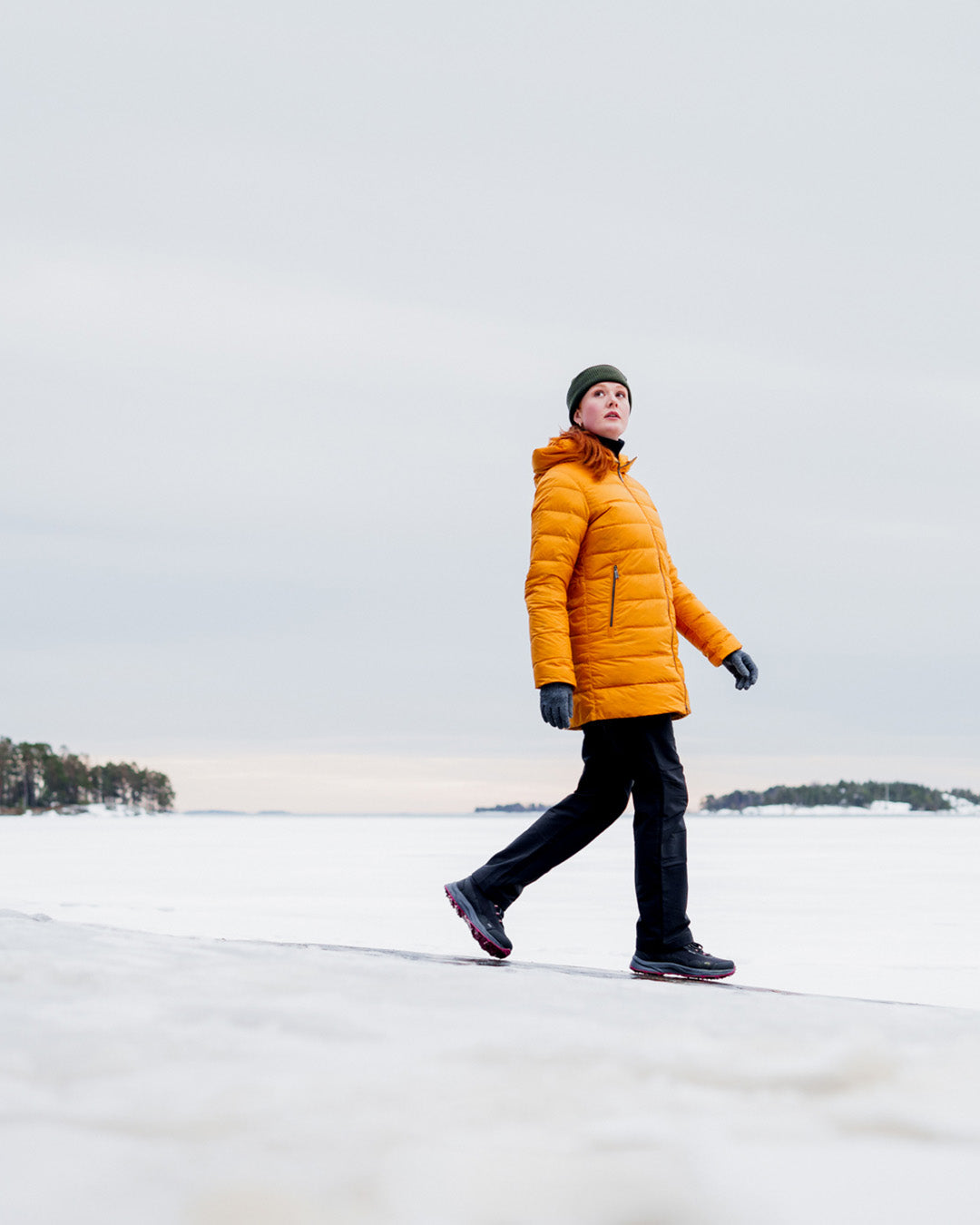 Nainen kävelee lumisella jäällä yllään vaakatikattu, keltainen Ruut-kevytuntuvatakki (väri: meripihka), mustat housut ja vihreä pipo. Taustalla näkyy metsää ja horisontissa saaria.