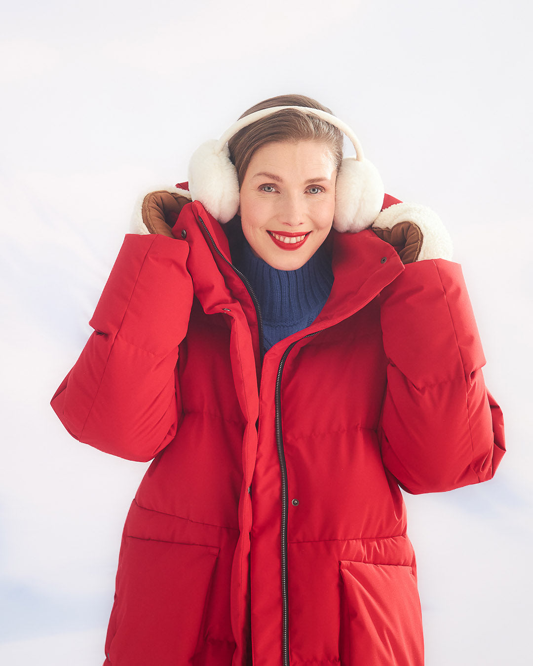 Nainen pitää käsillään punaisen (väri:chili) Siiri-untuvaparkatakin hupusta kiinni. Takissa on etupuolella suuret taskut ja vetoketju. Hänellä on päässään suuret valkoiset korvaläpät ja sininen pooloneule, taustalla on vaalea talvinen maisema.