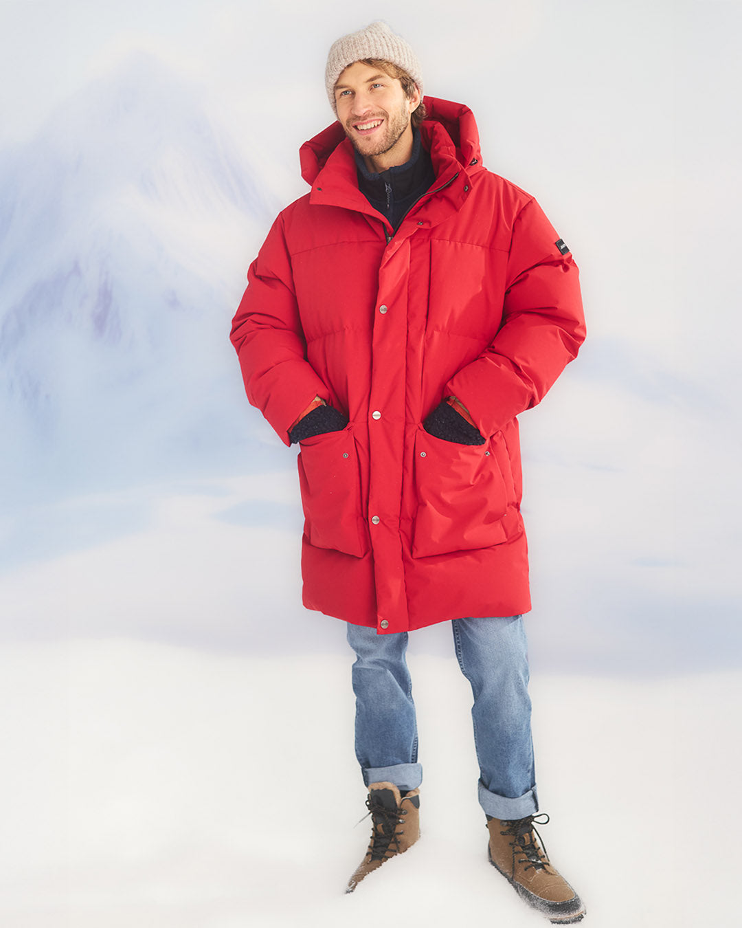 Mies seisoo lumisessa maisemassa päällään pitkä, punainen (väri: chili) Tuisku untuvaparkatakki, jossa on suuri huppu ja etutaskut. Hänellä on päällään siniset farkut, vaaleanharmaa neulepipo ja ruskeat nauhalliset talvikengät.