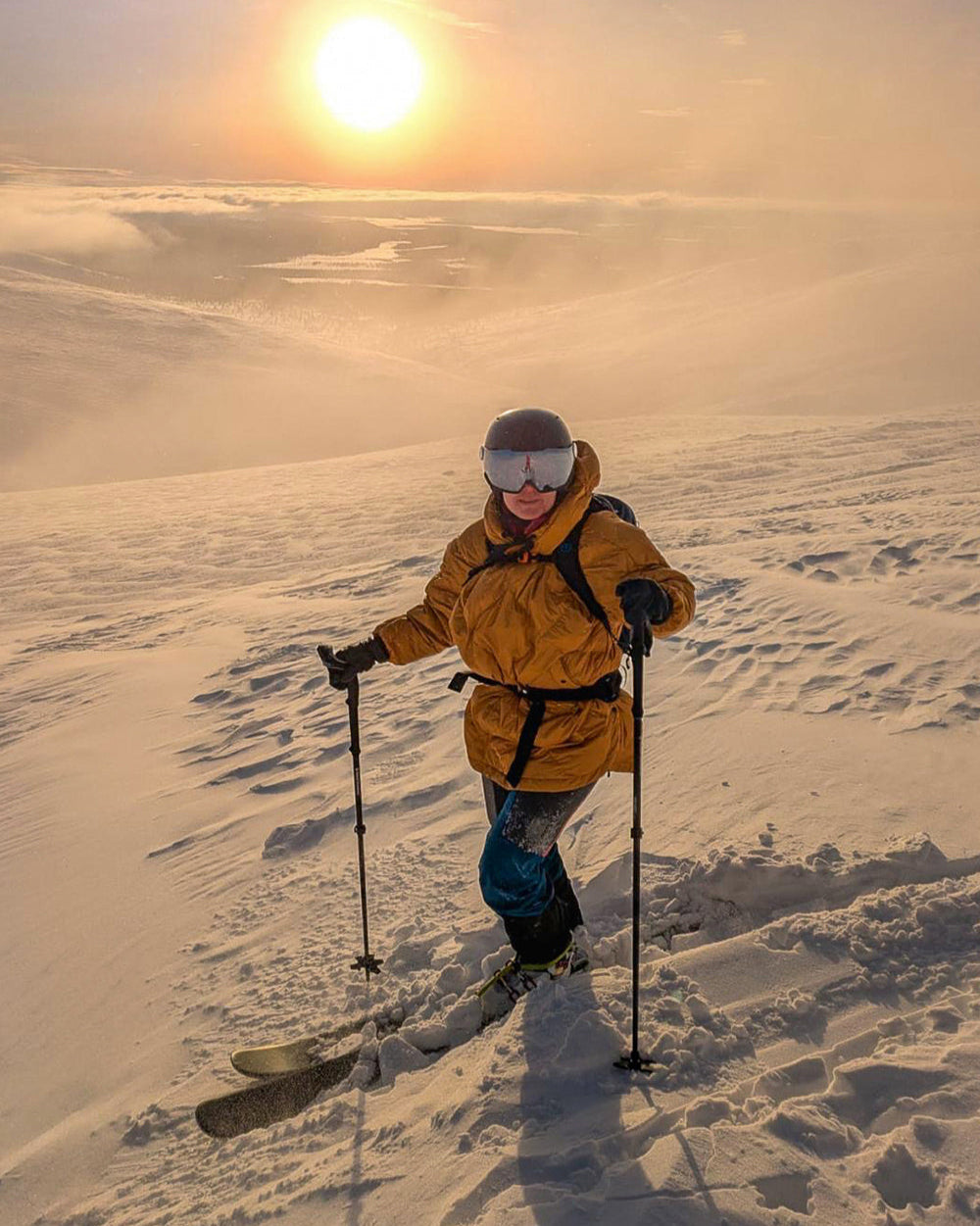 Hiihtäjä seisoo lumisella rinteellä auringonlaskussa yllään keltainen Velho-untuva-anorakki (väri: meripihka), jossa on huppu ja tikkaukset. Hänellä on laskettelulasit, sauvat ja sukset.