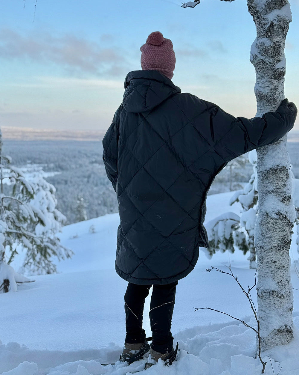 Henkilö seisoo lumisessa maisemassa selin kameraan yllään musta Velho-untuva-anorakki, jossa on timanttitikattu pinta ja huppu. Takki ulottuu polven yläpuolelle ja istuu väljästi.