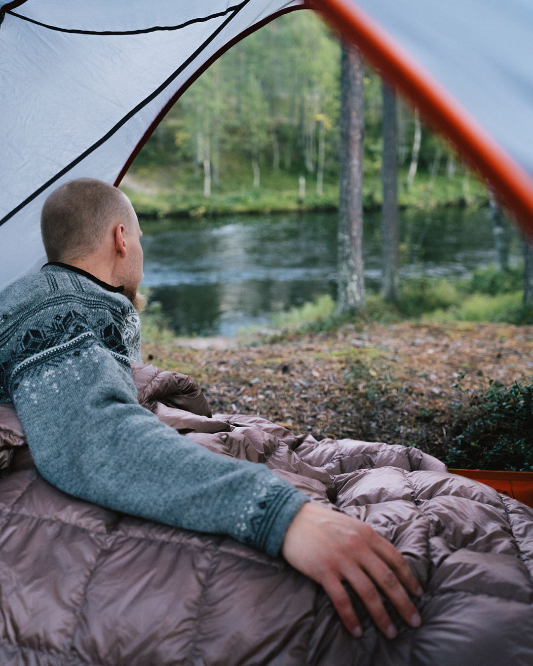 Henkilö makaa sinisessä villapaidassa harmaan Kulkuri-untuvatorkkupeiton alla teltassa ja katsoo joen suuntaan. Teltasta avautuu näkymä metsäiseen jokimaisemaan, jossa on sammalpohjaa ja puita joen rannalla.