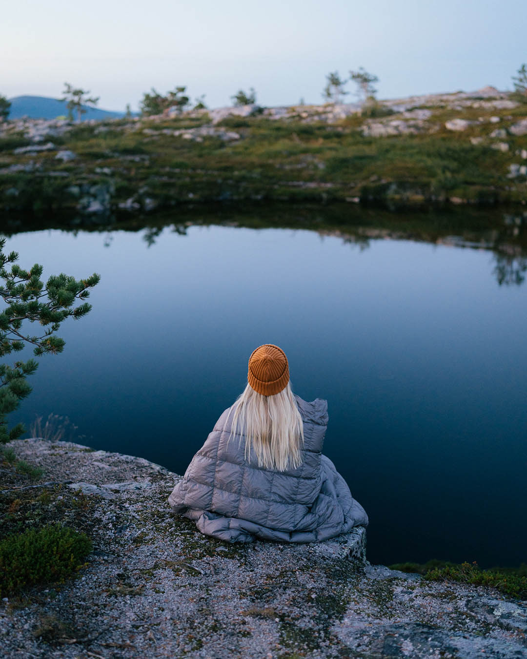 Henkilö istuu selin harmaan Kulkuri-untuvatorkkupeiton sisällä tyynen veden äärellä kallioisessa maisemassa. Hänellä on oranssi pipo ja pitkät vaaleat hiukset. Vedenpinta heijastaa sinertävää taivasta ja taustalla näkyy tunturikasvillisuutta.