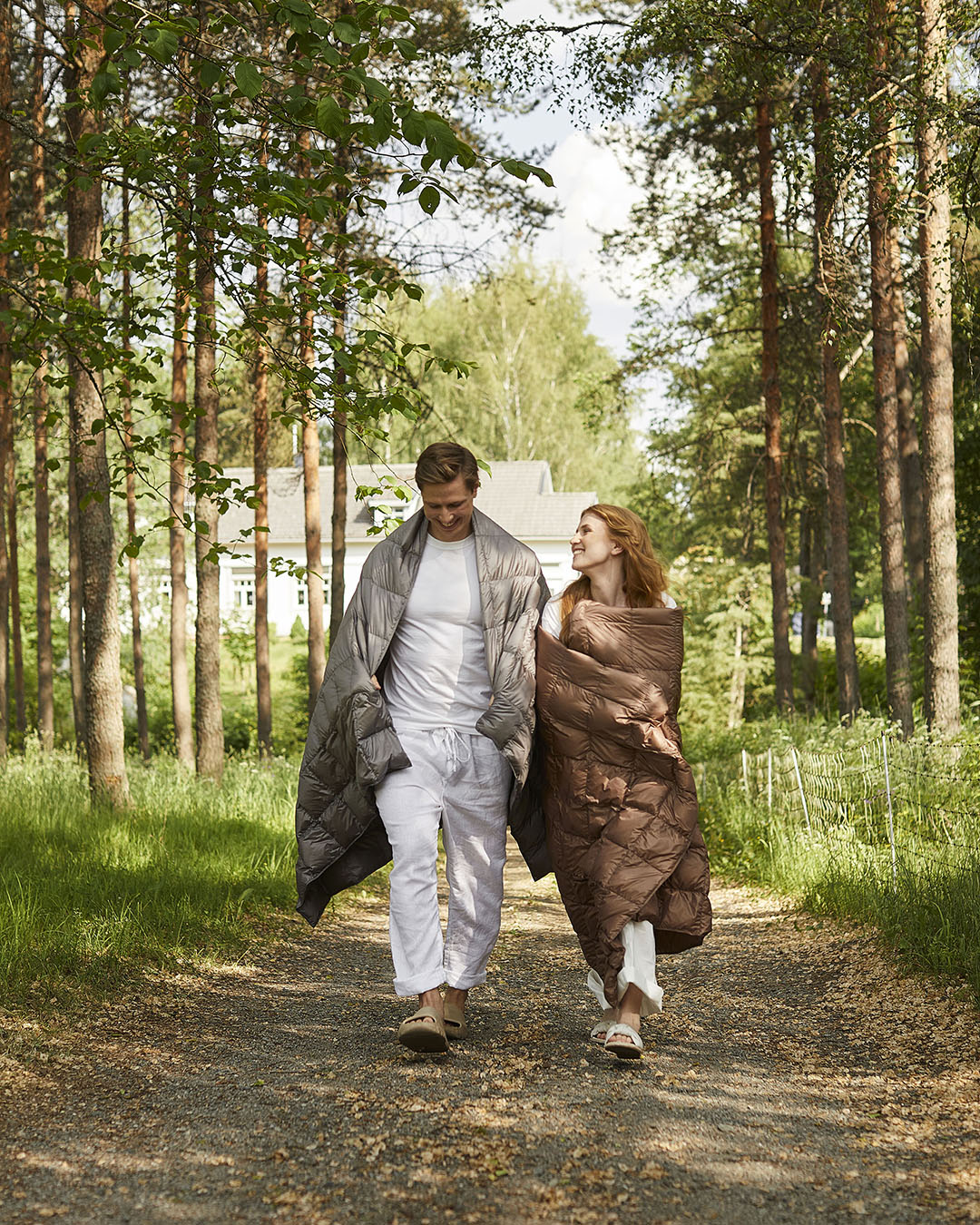 Kaksi henkilöä kävelee metsäistä hiekkatietä pitkin, molemmat kääriytyneinä Kulkuri-untuvatorkkupeittoihin – toisella harmaa ja toisella ruskea (väri: pähkinä). He katsovat toisiaan hymyillen.