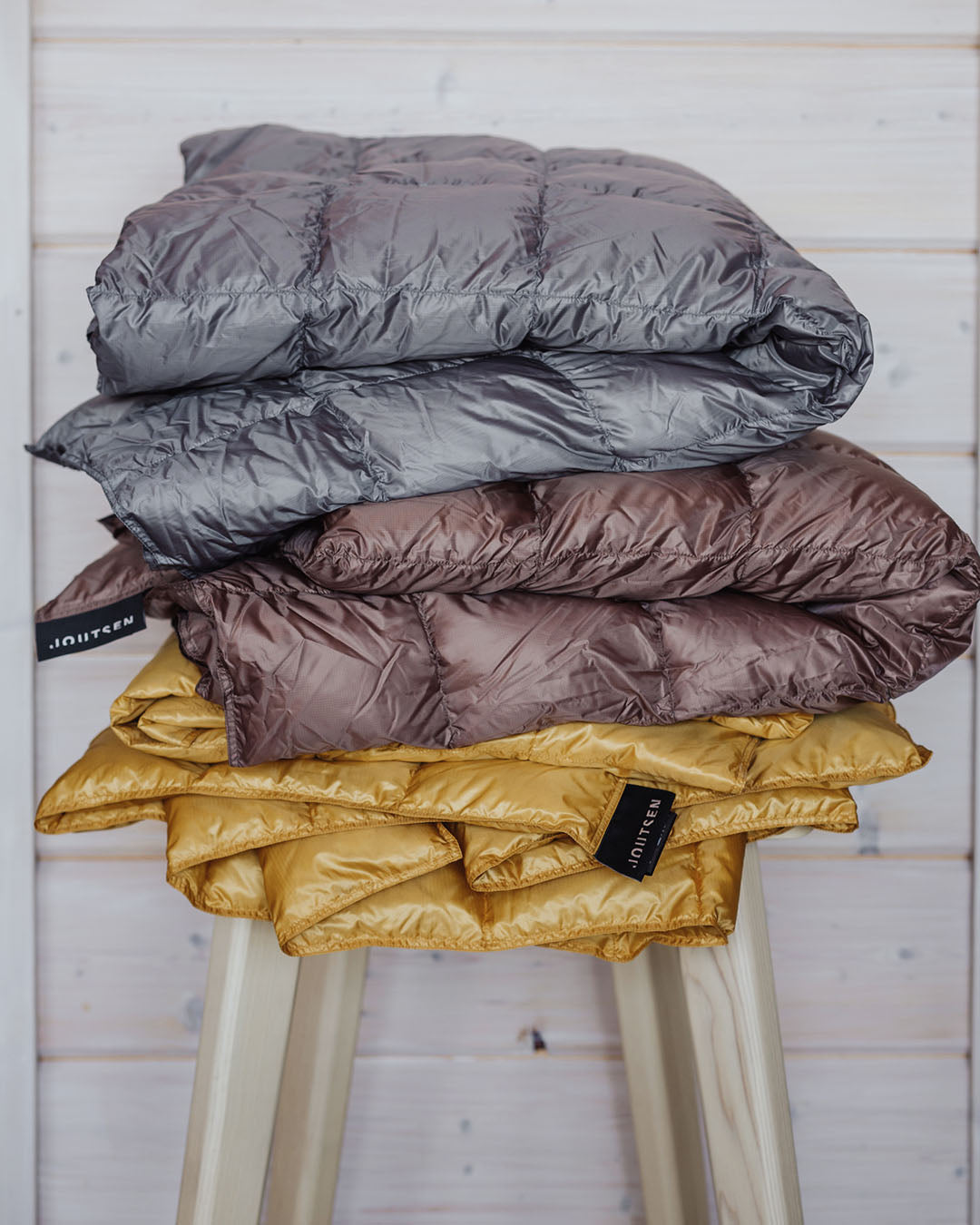 Kolme Kulkuri-untuvatorkkupeittoa on taiteltu päällekkäin vaalean puujakkaran päälle. Peitot ovat väriltään harmaa, ruskea (väri: pähkinä) ja keltainen (väri: Toscanan aurinko), ja niissä näkyy Joutsen-tuotemerkki. Taustalla vaalea vaakapaneeliseinä.