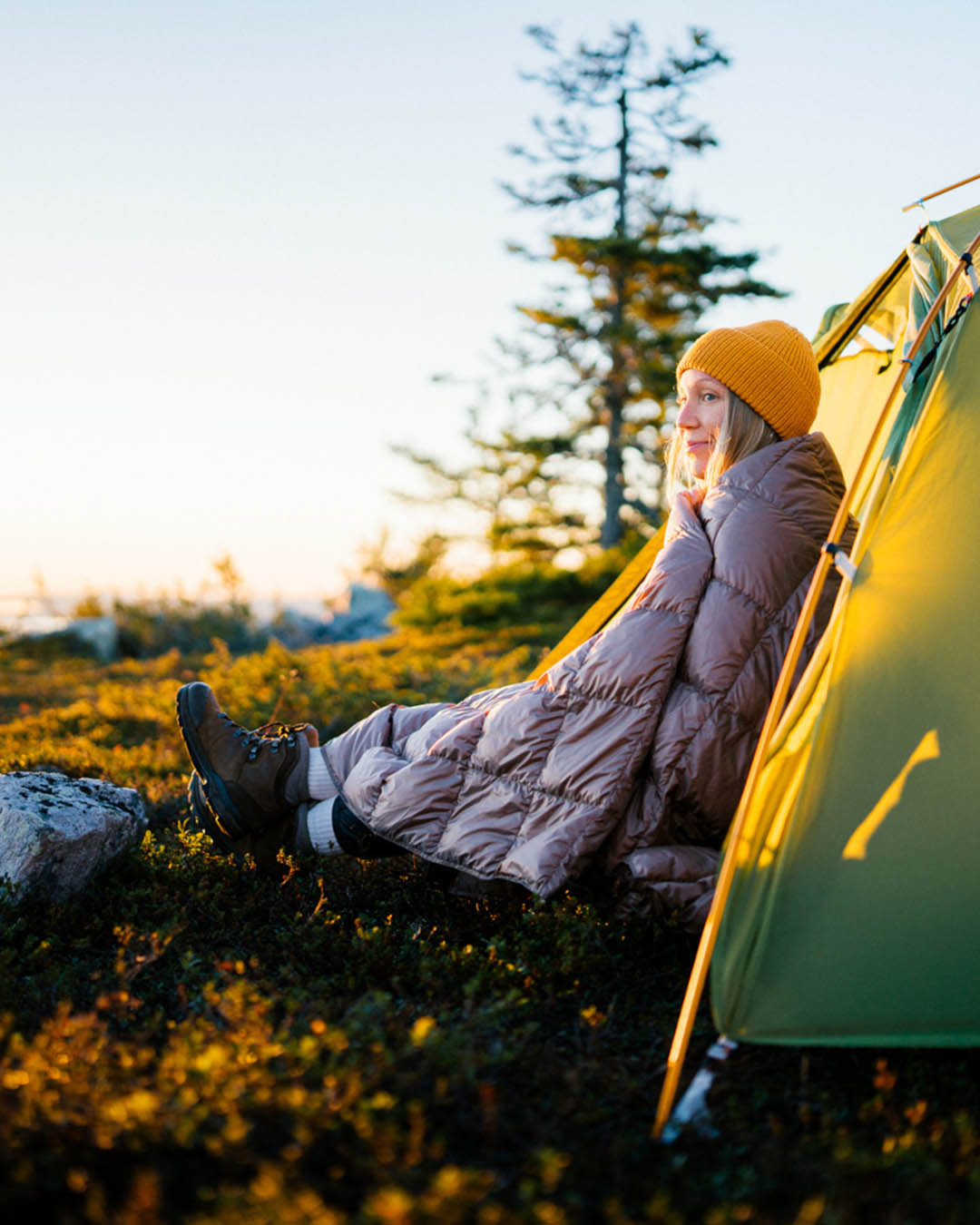 Henkilö istuu teltan edustalla luonnon keskellä aamuauringon valossa, kääriytyneenä harmaaseen Kulkuri-untuvatorkkupeittoon. Hänellä on keltainen pipo ja vaelluskengät, ja katse suuntautuu horisonttiin.