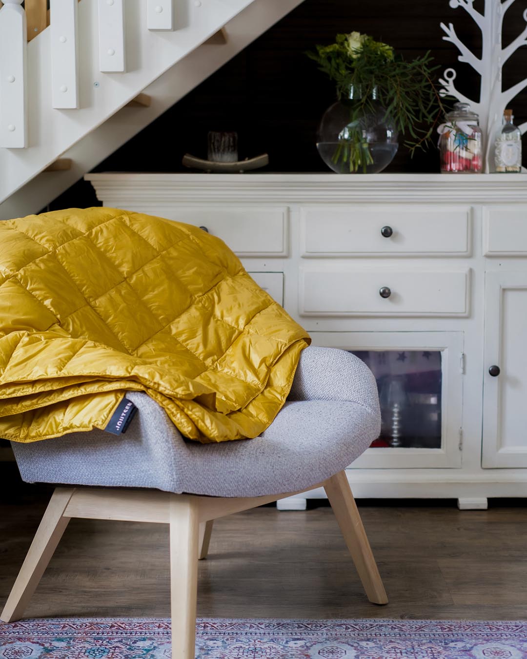 Keltainen Kulkuri-untuvatorkkupeitto on laskostettu vaaleanharmaan nojatuolin selkänojalle kodikkaassa sisätilassa. Taustalla valkoinen kaapisto, lasimaljakko vihreine oksineen ja koristeellinen valkoinen puufiguuri.