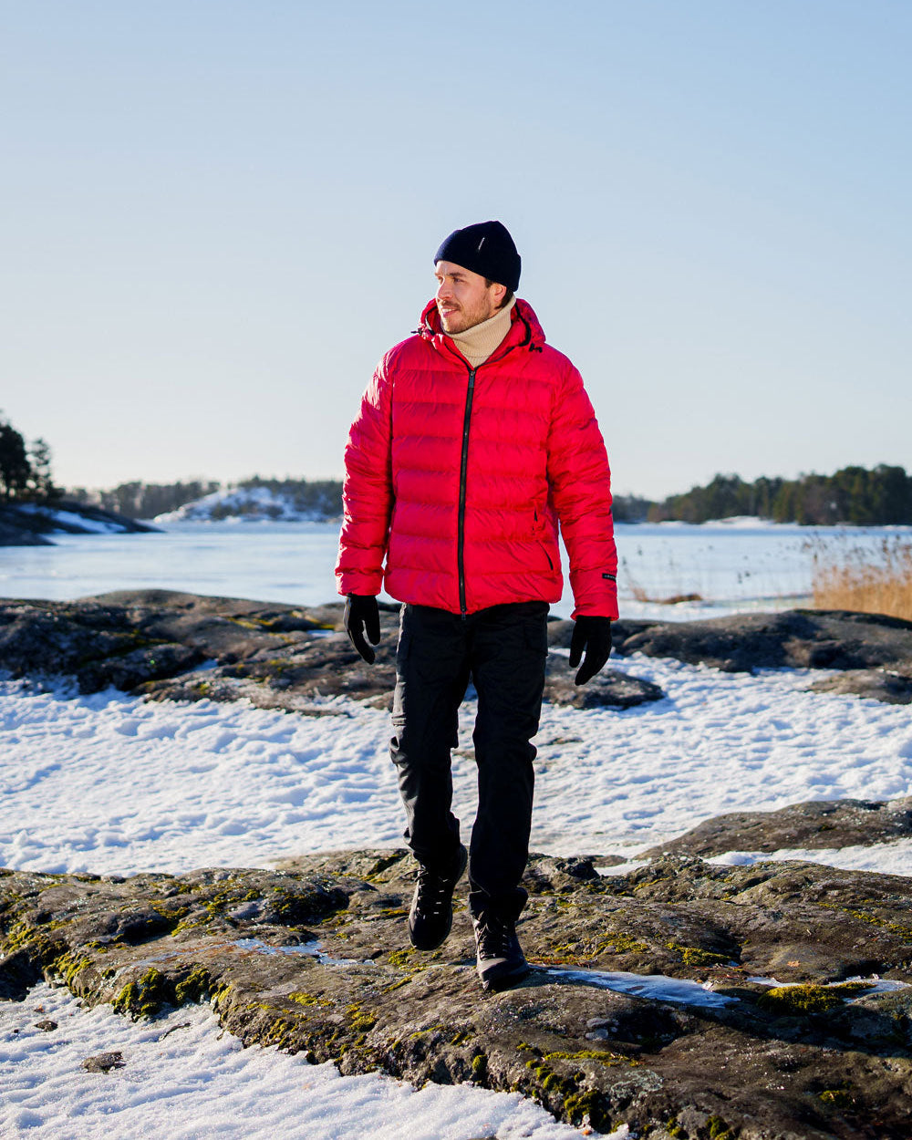 Mies kävelee lumisella kalliolla pukeutuneena vadelmanväriseen Kuulas-kevytuntuvatakkiin. Takissa on kiiltävä pinta, vaakasuuntaiset tikkaukset ja huppu. Asusteina musta pipo, hanskat ja housut.