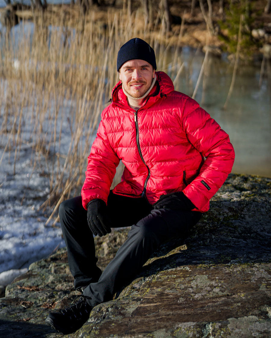 Henkilö istuu kallion päällä talvisessa maisemassa pukeutuneena vadelmanväriseen Kuulas-kevytuntuvatakkiin. Takki on lyhyt, kevyesti topattu ja siinä on kiiltävä pinta sekä huppu. Asukokonaisuuteen kuuluu myös musta pipo, hanskat ja housut.