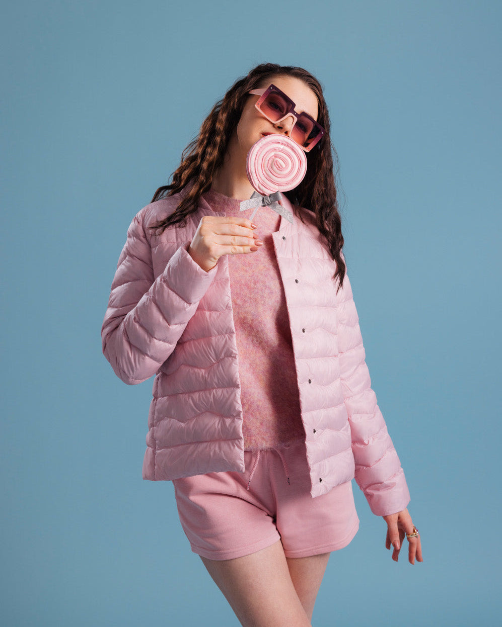 Nainen pukeutuneena vaaleanpunaiseen Lootus-kevytuntuvatakkiin (väri: mansikkavaahto), sävyyn sopivaan neulepaitaan ja shortseihin. Asuun kuuluu myös suuret aurinkolasit ja pyöreä tikkari.