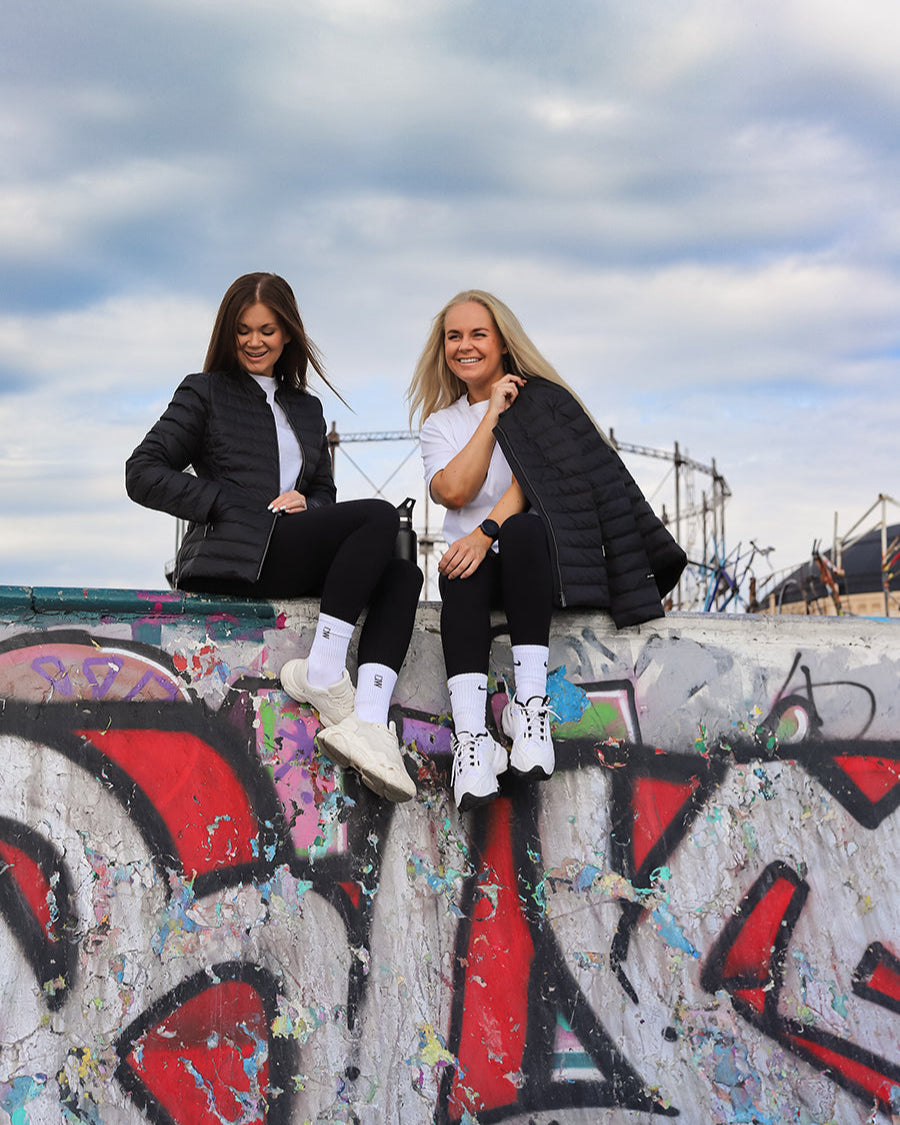 Kaksi naista istuu graffitiseinällä pukeutuneina mustiin Maana-kevytuntuvatakkeihin. Molemmilla on takkien alla valkoiset paidat, mustat leggingsit ja valkoiset lenkkarit.