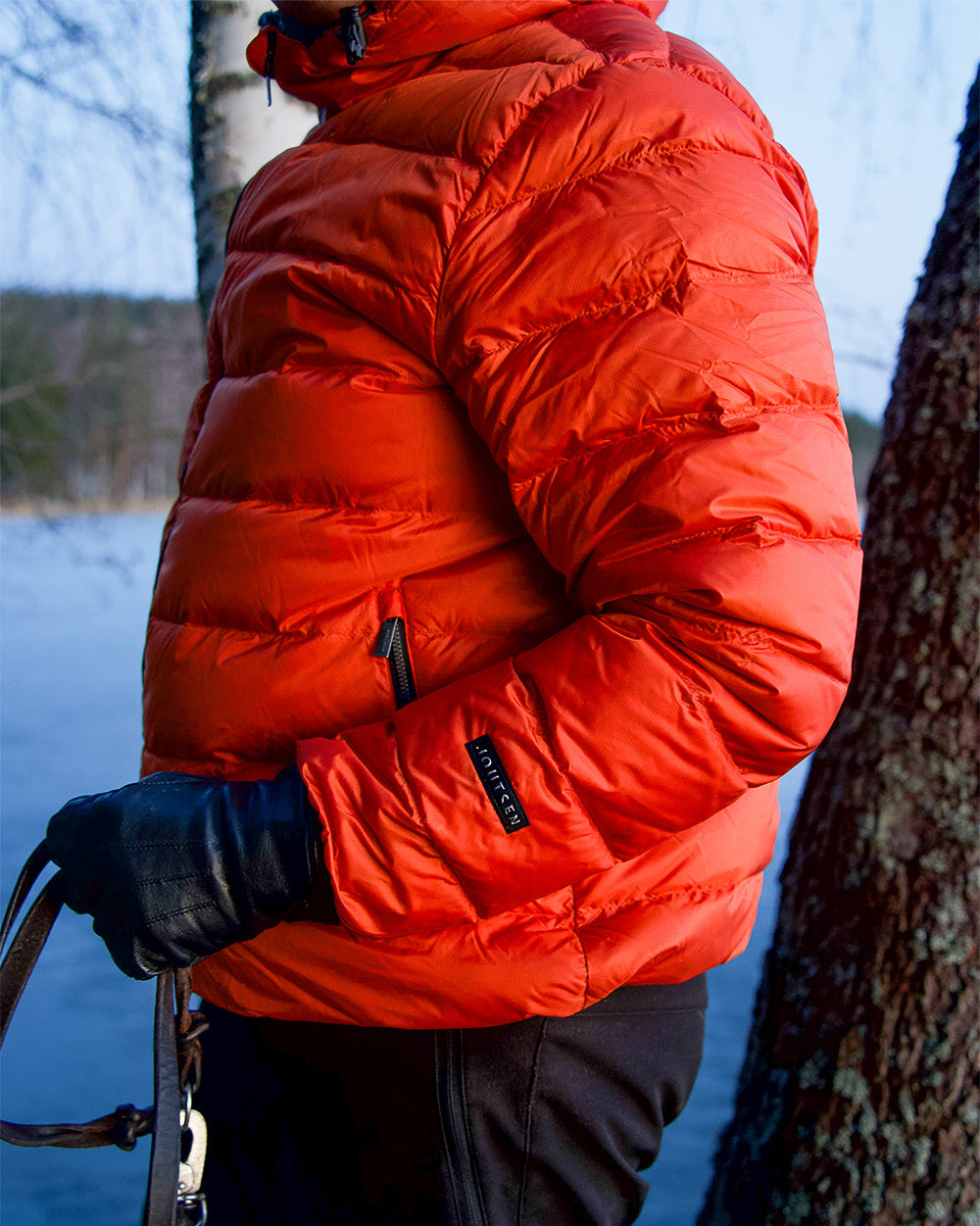 Henkilö seisoo ulkona järven rannalla yllään oranssi Ruska-kevytuntuvatakki (väri: syksyn oranssi), jossa on vaakasuuntaiset tikkaukset ja vetoketjutaskut. Hihassa on Joutsen-logomerkki.