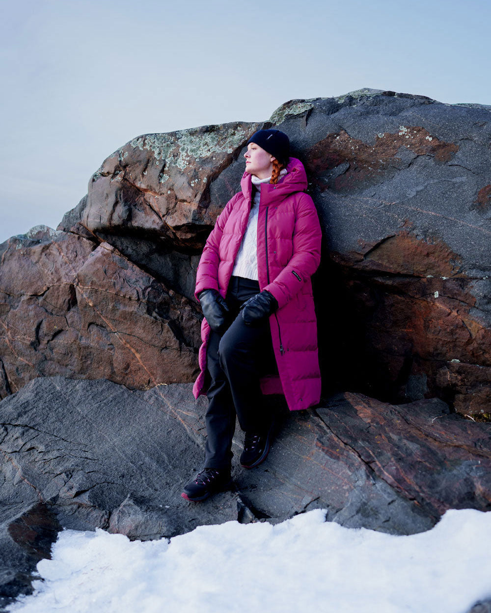 Nainen seisoo lumisella rannalla suuren kallion edessä yllään fuksianvärinen pitkä untuvatakki (väri: pioni), jossa on huppu. Asuun kuuluu myös tummat housut, mustat kengät, mustat nahkahanskat ja tumma pipo.