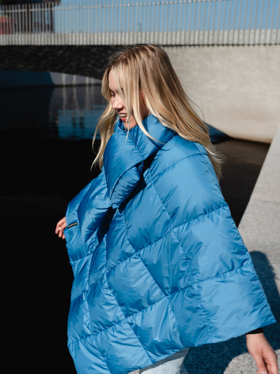 Hymyilevä henkilö ulkona auringonpaisteessa yllään kirkkaan sininen, pehmeä ja paksu Hiutale-untuvahuivi (väri: sinisiipi), joka on kiedottu hartioille. Alla hänellä on samanvärinen untuvaponcho.