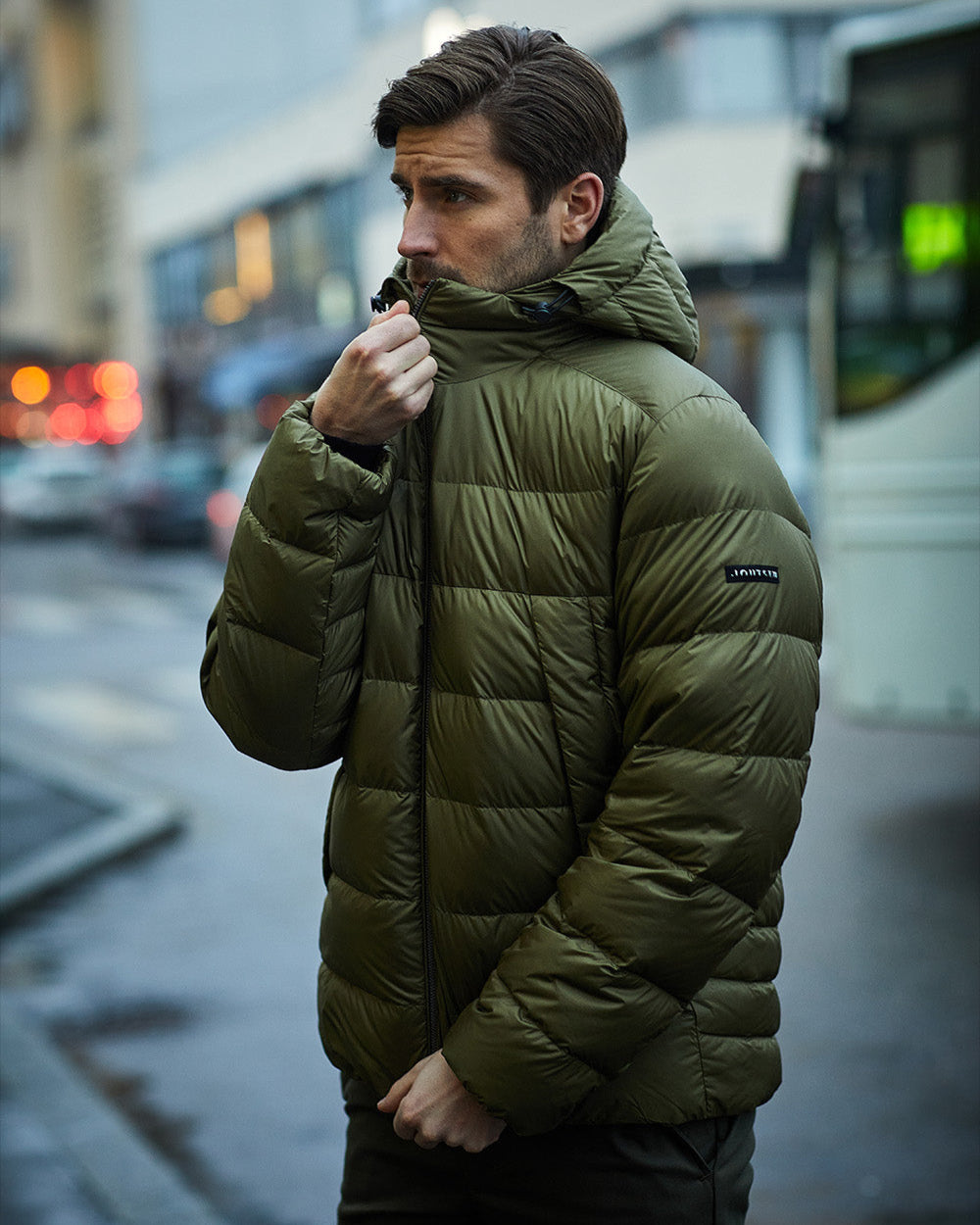 Mies ulkona kaupunkiympäristössä yllään oliivinvihreä Valto-kevytuntuvatakki (väri: kultainen oliivi), jossa on vaakasuorat tikkaukset ja huppu.