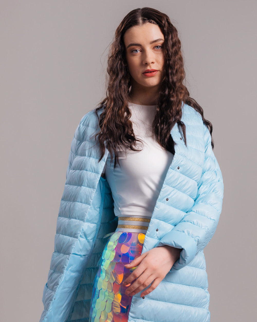 Nainen pukeutuneena vaaleansiniseen pidempään Vuokko-kevytuntuvatakkiin (väri: jääkaramelli), valkoiseen yläosaan ja moniväriseen, paljeteilla koristeltuun alaosaan. Takki on auki.