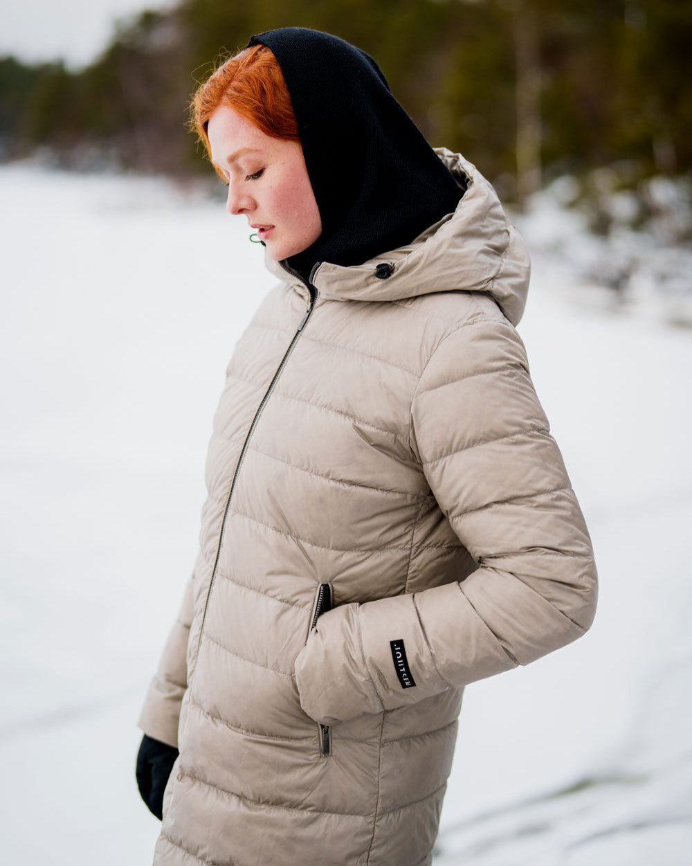 Nainen seisoo lumisessa maisemassa ja katsoo alaviistoon. Hänellä on yllään pitkä, tikattu Hanne-kevytuntuvatakki, jossa on huppu ja vetoketjukiinnitys. Takki on vaalea beige (väri: samppanja), ja sen sivuilla on vetoketjutaskut. Naisen päässä on musta huppu ja hänellä on mustat hansikkaat.