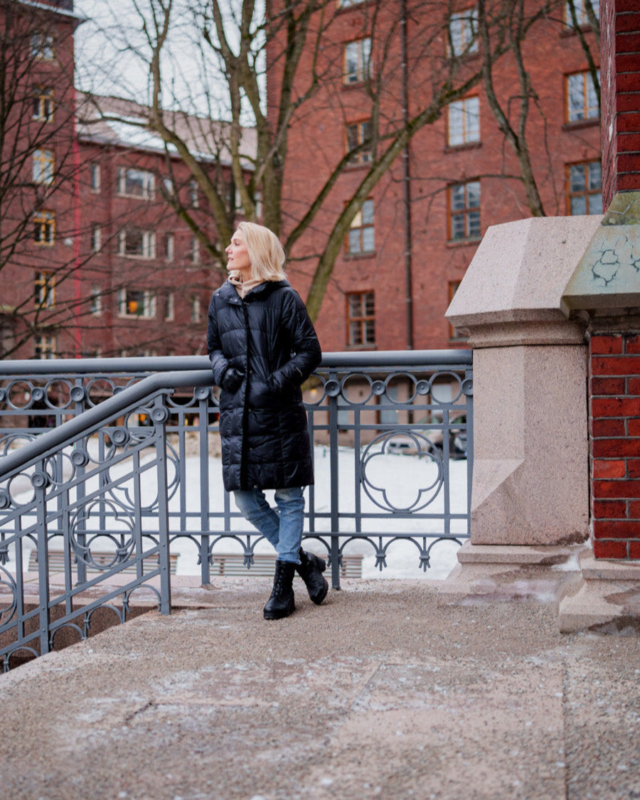 Nainen seisoo ulkona talvisessa kaupunkiympäristössä pukeutuneena mustaan, pitkään ja kapealinjaiseen Essi-kevytuntuvatakkiin (väri: hiilenmusta), jossa on huppu. Asuun kuuluu myös siniset farkut ja mustat nauhalliset kengät. Taustalla tiilirakennuksia ja luminen maa.