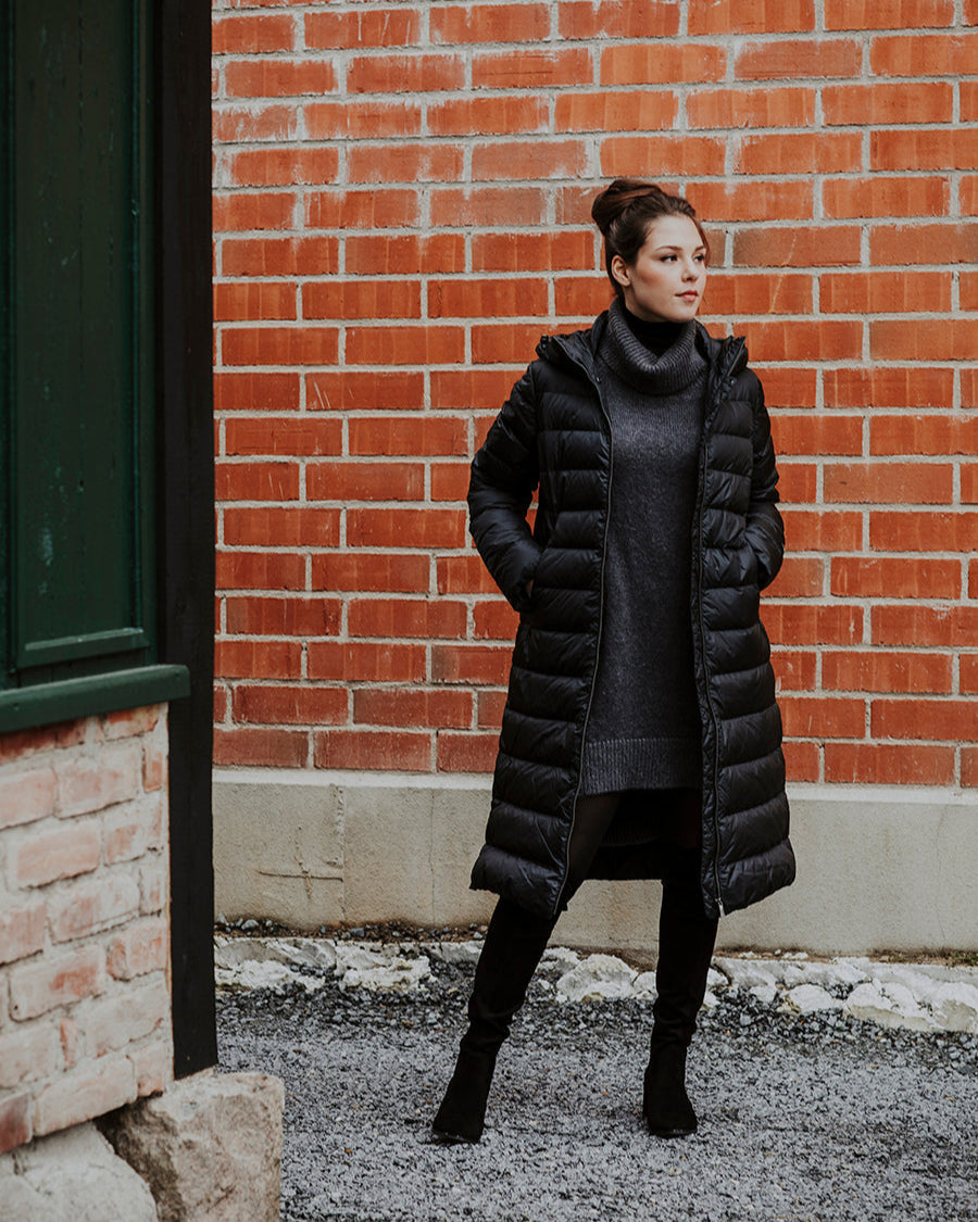 Nainen seisoo ulkona tiiliseinän edessä kädet kevyesti Hanne-kevytuntuvatakin taskuissa. Hänellä on yllään pitkä, tikattu kevytuntuvatakki, jossa on huppu ja vetoketjukiinnitys. Takki on musta (väri: hiilenmusta) ja sen alle on puettu pitkä neulemekko ja mustat saappaat.