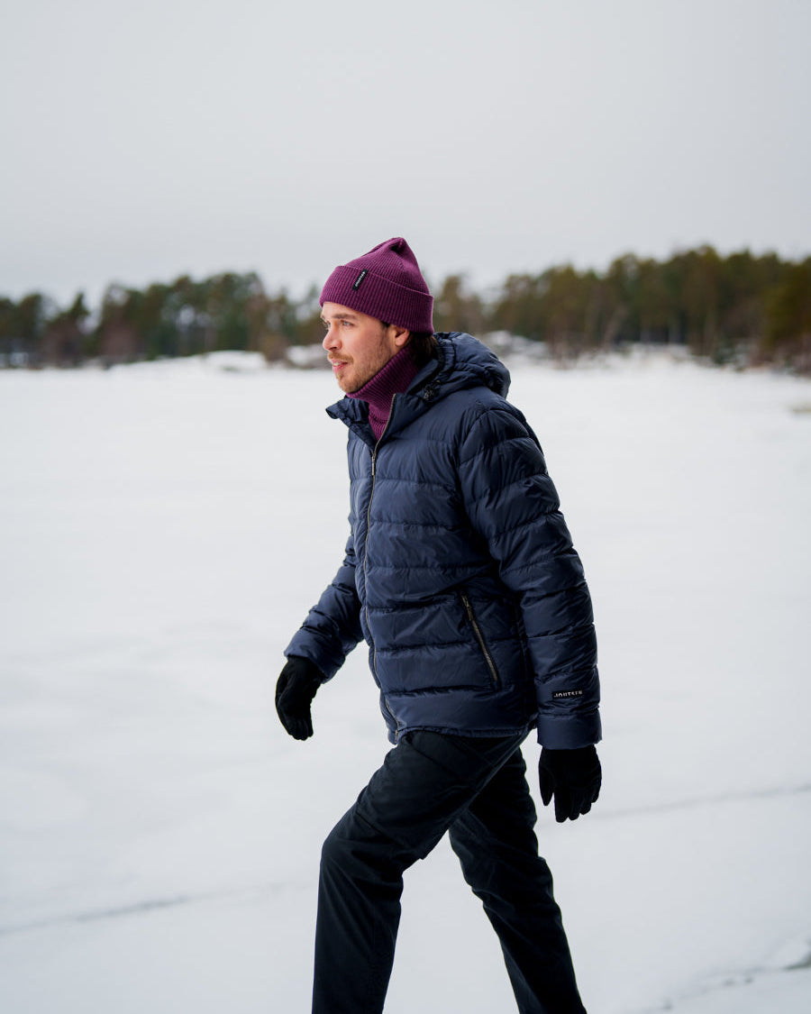 Henkilö kävelee lumisessa maisemassa yllään tummansininen Ruska-kevytuntuvatakki (väri: pohjoisen sininen), jossa on huppu ja vetoketjulliset taskut. Päässä hänellä on violetti pipo.