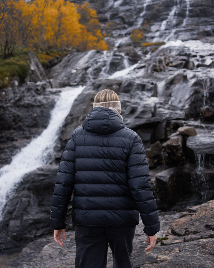 Henkilö seisoo selin vesiputouksen edessä yllään musta Ruska-kevytuntuvatakki (väri: pohjoisen musta), jossa on huppu ja vaakasuuntaiset tikkaukset.