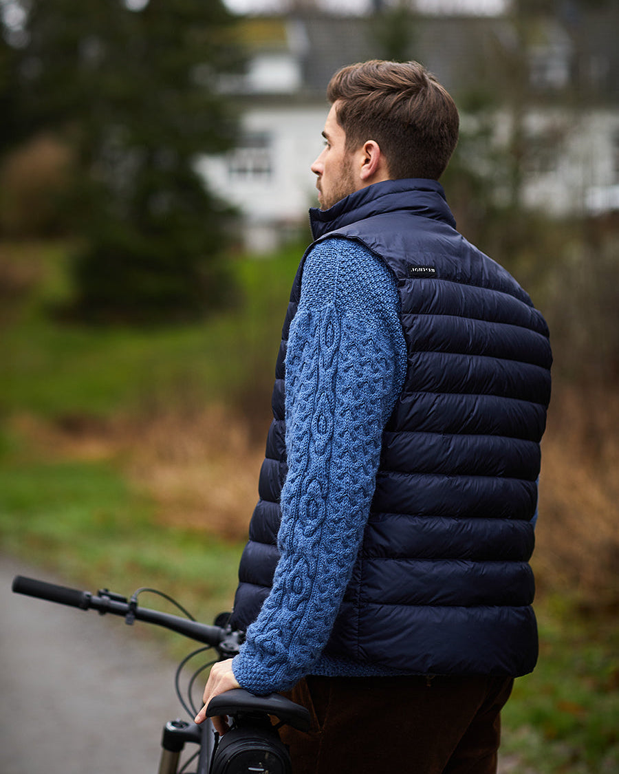 Mies seisoo polkupyörän kanssa ulkona yllään tummansininen Pouta-untuvaliivi (väri: yönsininen), jossa on leveät vaakasuuntaiset tikkaukset ja pystykaulus.
