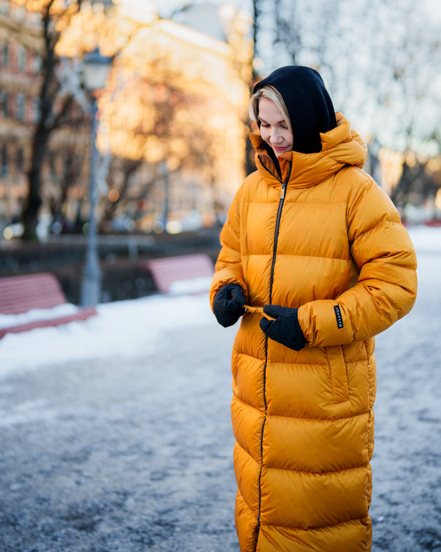 Nainen seisoo talvisella kadulla pukeutuneena meripihkan väriseen Estrid-untuvatakkiin, mustiin hanskoihin ja mustaan huppuun. Hän hymyilee ja pitää kiinni takin vyötäröllä olevista kiristysnaruista. Taustalla on penkkejä, lumipeitettä ja kaupunkimaisema.