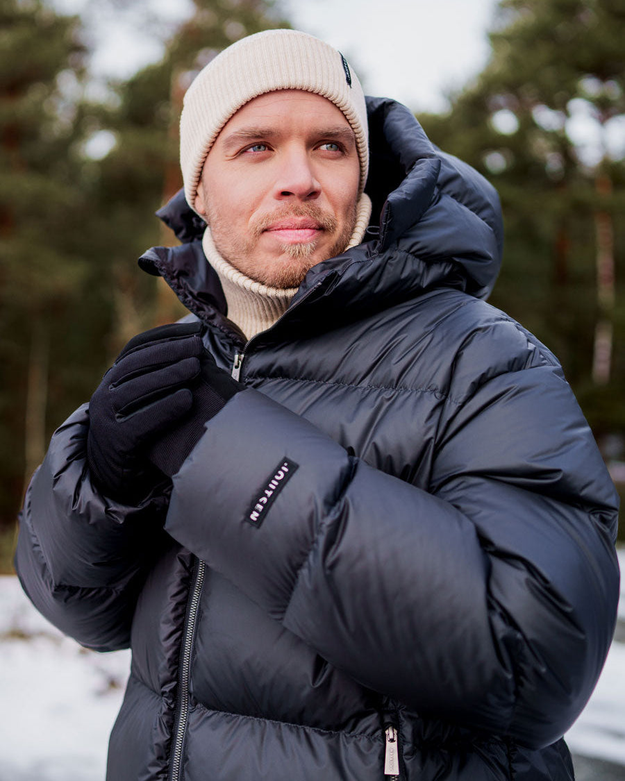 Mies pukeutuneena hiilenmustaan Knut-untuvatakkiin, jonka pinta on hieman kiiltävä ja vaakatikattu. Hihassa on musta Joutsen-logo. Hänellä on beige pipo ja vaalea poolokauluspaita sekä mustat hanskat. Taustalla näkyy talvinen metsämaisema.