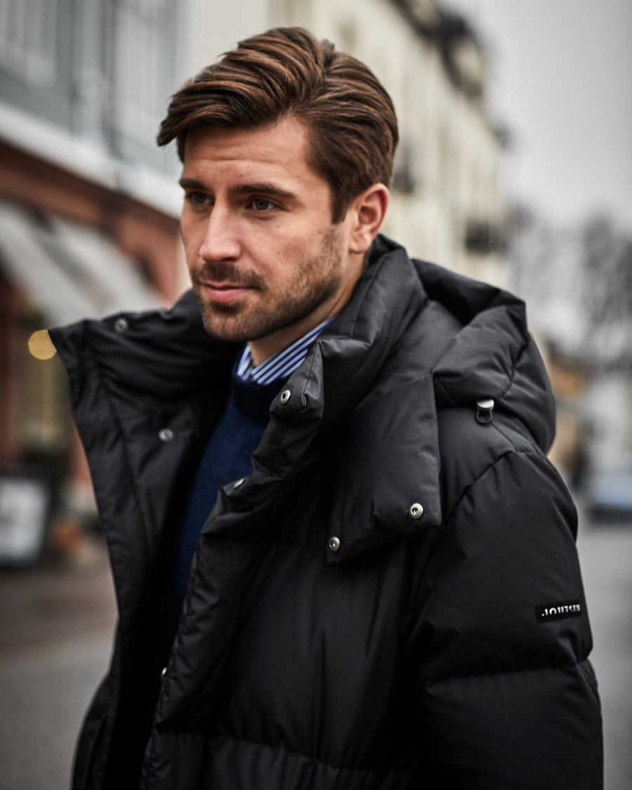 Mies pukeutuneena mustaan, lakritsinväriseen Koski-untuvatakkiin, jossa on mattapinta, leveät tikkaukset ja irrotettava huppu. Olkavarressa on Joutsen-logo. Takki on yhdistetty kauluspaitaan ja neuleeseen.