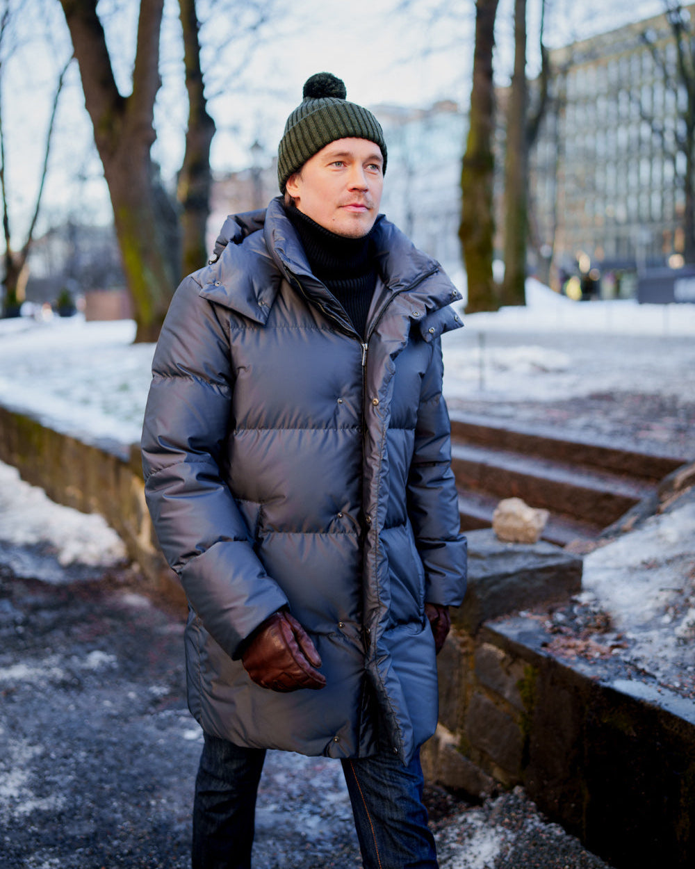 Mies kävelee talvisessa puistossa pukeutuneena laavanväriseen Koski-untuvatakkiin, jossa on irrotettava huppu. Asukokonaisuuteen kuuluu myös vihreä tupsupipo, musta pooloneule, farkut ja ruskeat nahkahanskat.