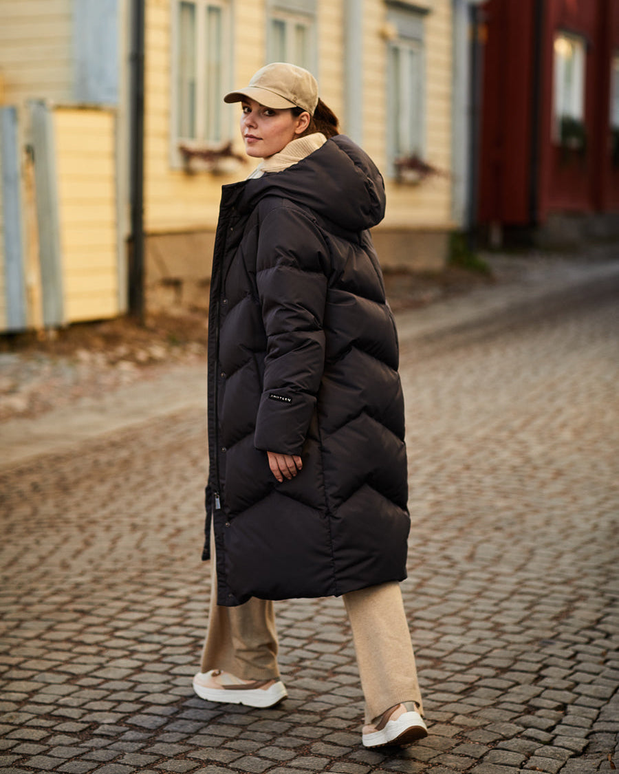 Nainen kävelee katsoen takaviistoon mukulakivikadulla yllään musta, pitkä ja paksu Reina-untuvatakki (väri: mustatorvisieni), beige lippalakki, beiget housut ja vaaleat lenkkarit.