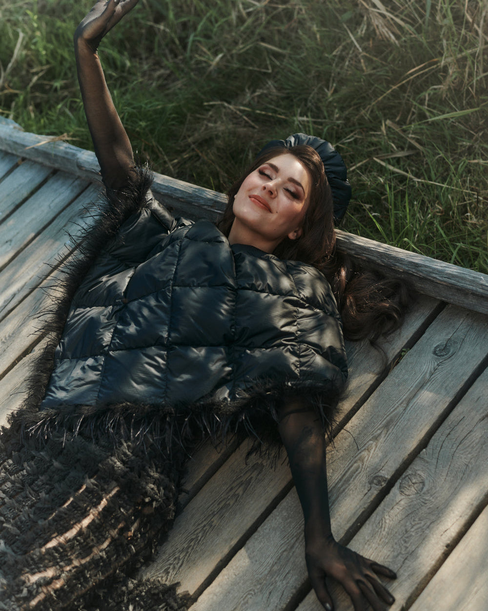 Nainen makaa puisella laiturilla yllään musta, kiiltäväpintainen Lumous-untuvaponcho ja siihen sointuva Pumpuli-untuvabaskeri. Ponchossa on vinot tikkaukset ja kevyt höyhenkoristelu helmassa.