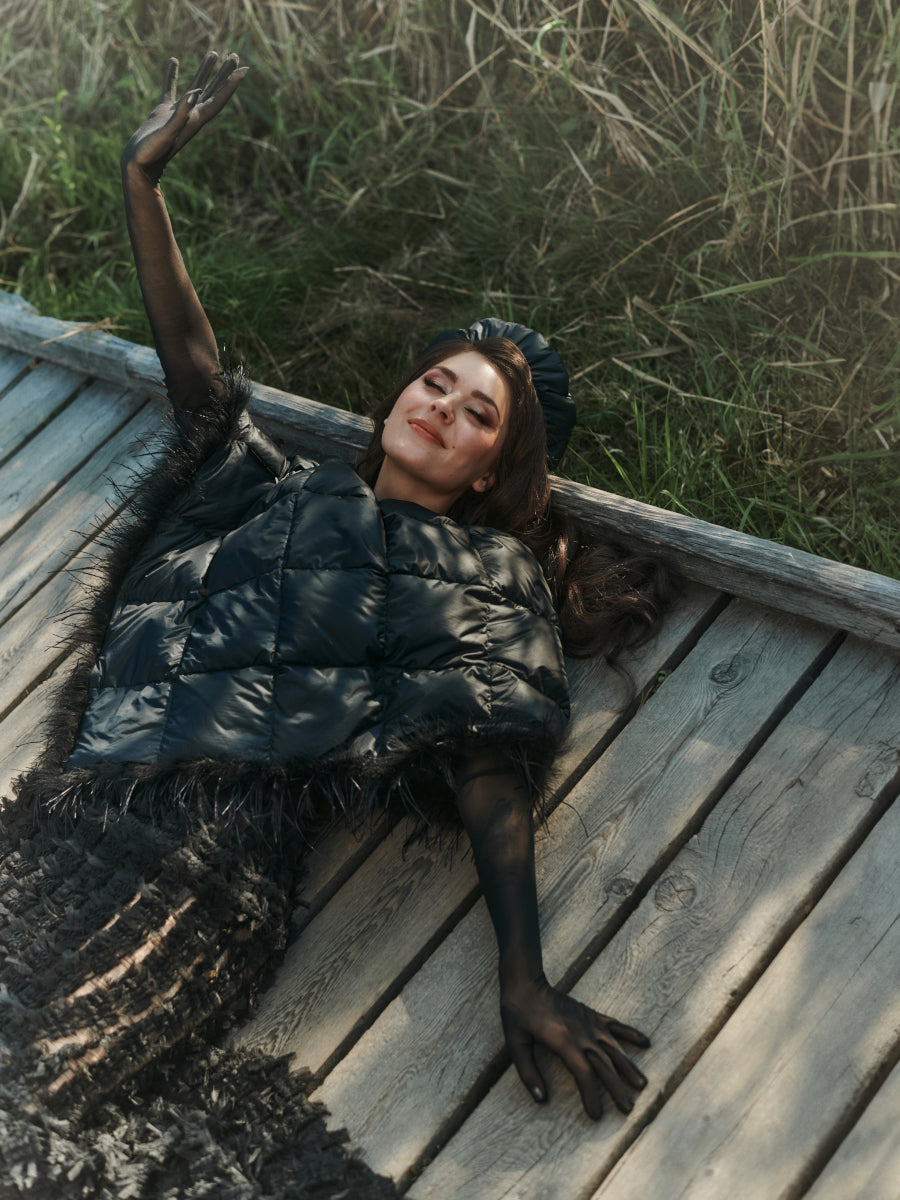 Henkilö makaa puisella laiturilla yllään musta, kiiltäväpintainen Pumpuli-untuvabaskeri ja samansävyinen asukokonaisuus, johon kuuluu musta höyhenkoristeinen Lumous-untuvaponcho ja läpikuultavat hihat.