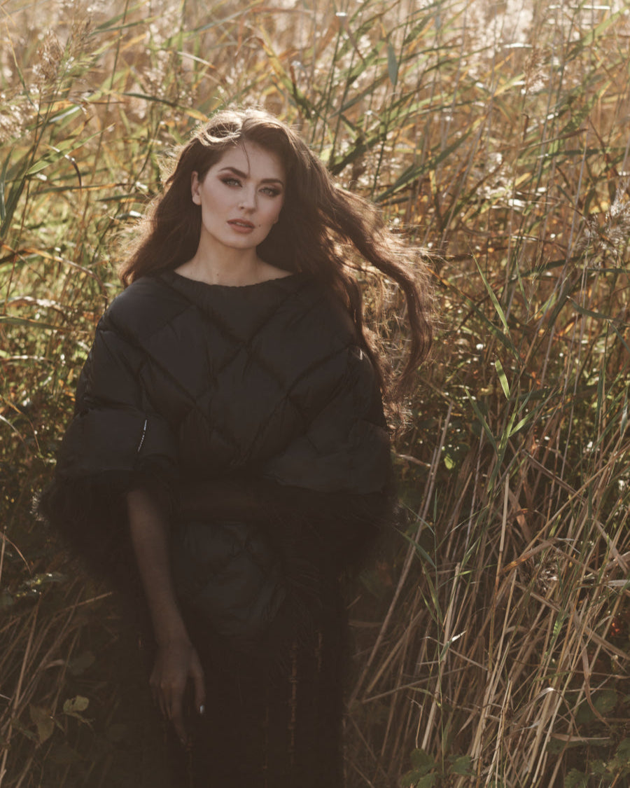 Nainen seisoo korkeassa heinikkossa yllään musta Lumous-untuvaponcho, jossa on kiiltävä pinta, vinot timanttikuviot ja helmassa kevyt höyhenkoristelu. Hänen hiuksensa hulmuavat kevyessä tuulessa.