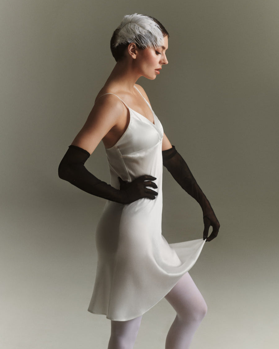 Nainen poseeraa neutraalissa studiovalossa yllään valkoinen Melodia-silkkimekko, jossa on ohuet olkaimet. Asuun on yhdistetty mustat pitkät hansikkaat, valkoiset sukkahousut ja höyhenkoriste hiuksissa.