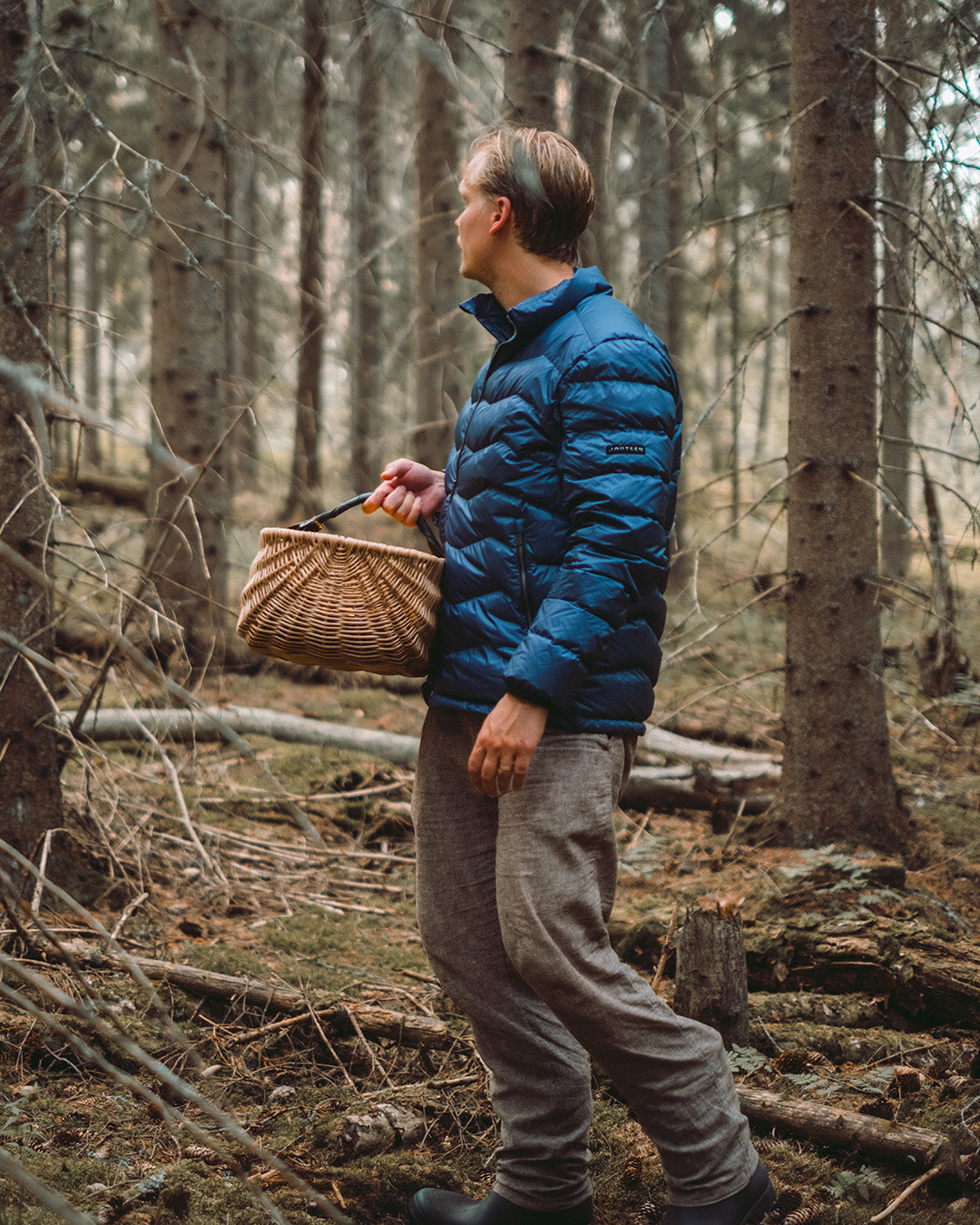 Mies seisoo metsässä pitäen kädessään punottua koria. Hänellä on yllään sininen Hile-kevytuntuvatakki (väri: myrskyluoto).