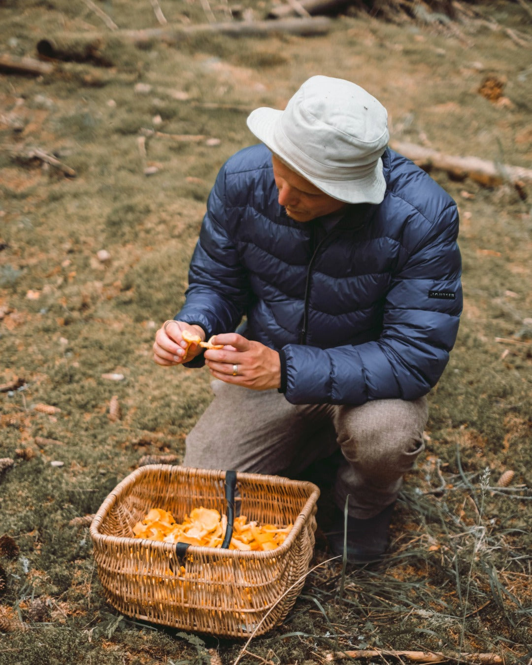 Mies on kyykistyneenä metsässä ja puhdistaa kantarelleja. Hänellä on yllään sininen Hile-kevytuntuvatakki (väri: myrskyluoto) ja vaalea lierihattu, edessään täyttymässä oleva sienikori.