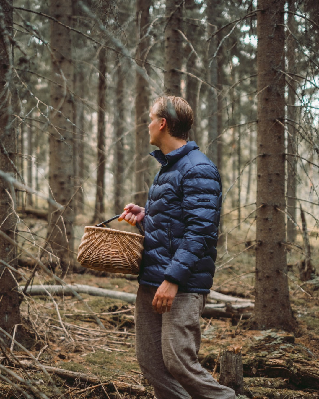 Mies seisoo metsässä pitäen kädessään punottua koria. Hänellä on yllään sininen Hile-kevytuntuvatakki (väri: myrskyluoto).