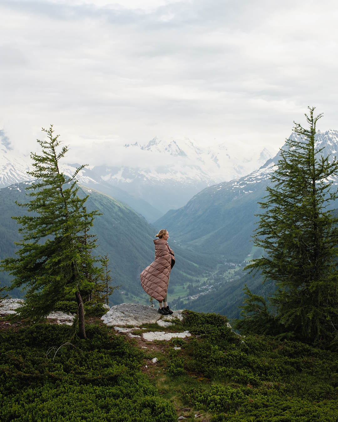 Henkilö seisoo vuorenrinteellä kahden havupuun välissä katsellen laaksoon. Hän on pukeutunut vaaleanruskeaan Kulkuri-untuvatorkkupeittoon (väri: pähkinä), joka hulmuaa tuulessa. Taustalla näkyvät vehreät vuoristomaisemat ja lumihuippuiset vuoret pilvien peittämää taivasta vasten.