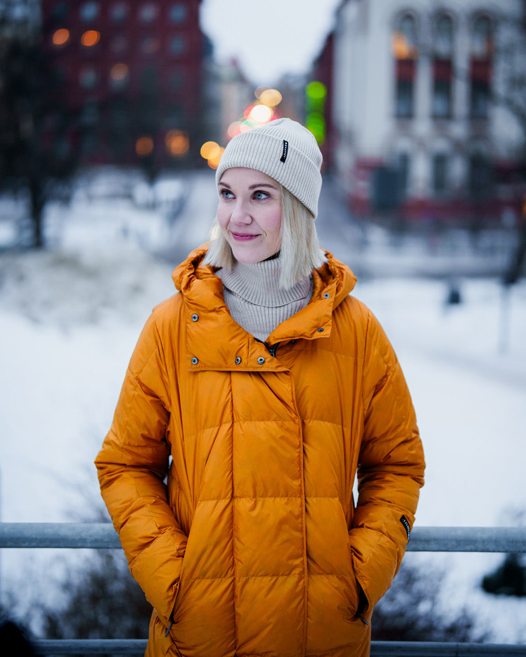 Nainen seisoo talvisessa kaupunkiympäristössä pukeutuneena keltaiseen, pitkään Essi-kevytuntuvatakkiin (väri: meripihka), jossa on huppu ja neliömäiset tikkaukset. Asuun kuuluu myös vaalea pipo ja poolokauluksinen neule. Taustalla näkyy luminen maisema ja rakennuksia.