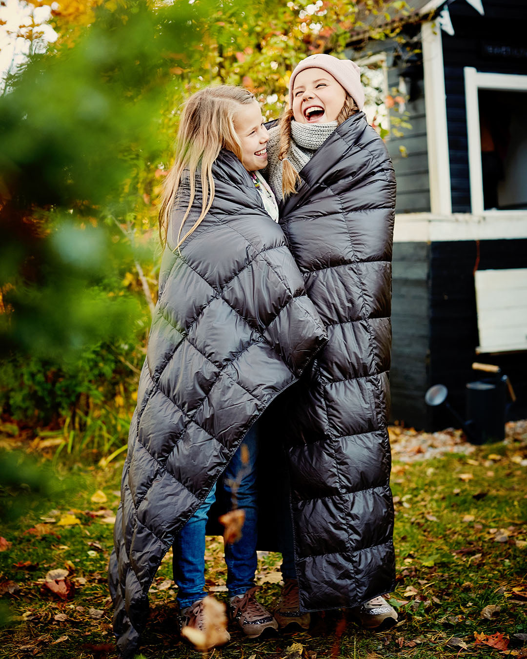 Kaksi hymyilevää lasta seisoo ulkona syksyisessä pihapiirissä kääriytyneenä tummanharmaan Kulkuri-untuvatorkkupeiton (väri: antrasiitti) sisään. He katsovat toisiaan nauraen, ja taustalla näkyy mökki sekä ruskan sävyjä puissa.