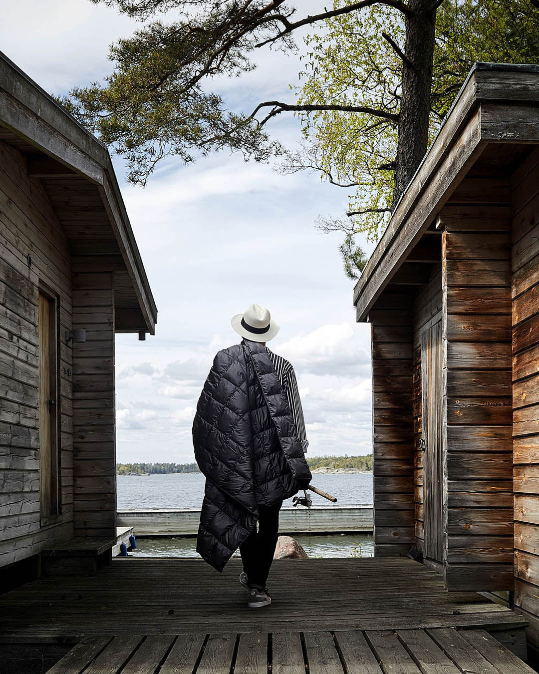 Henkilö kävelee laiturille kahden hirsisaunan välissä tumman harmaa Kulkuri-untuvatorkkupeitto (väri: antrasiitti) olallaan. Taustalla näkyy järvimaisema, puu ja pilvinen taivas.