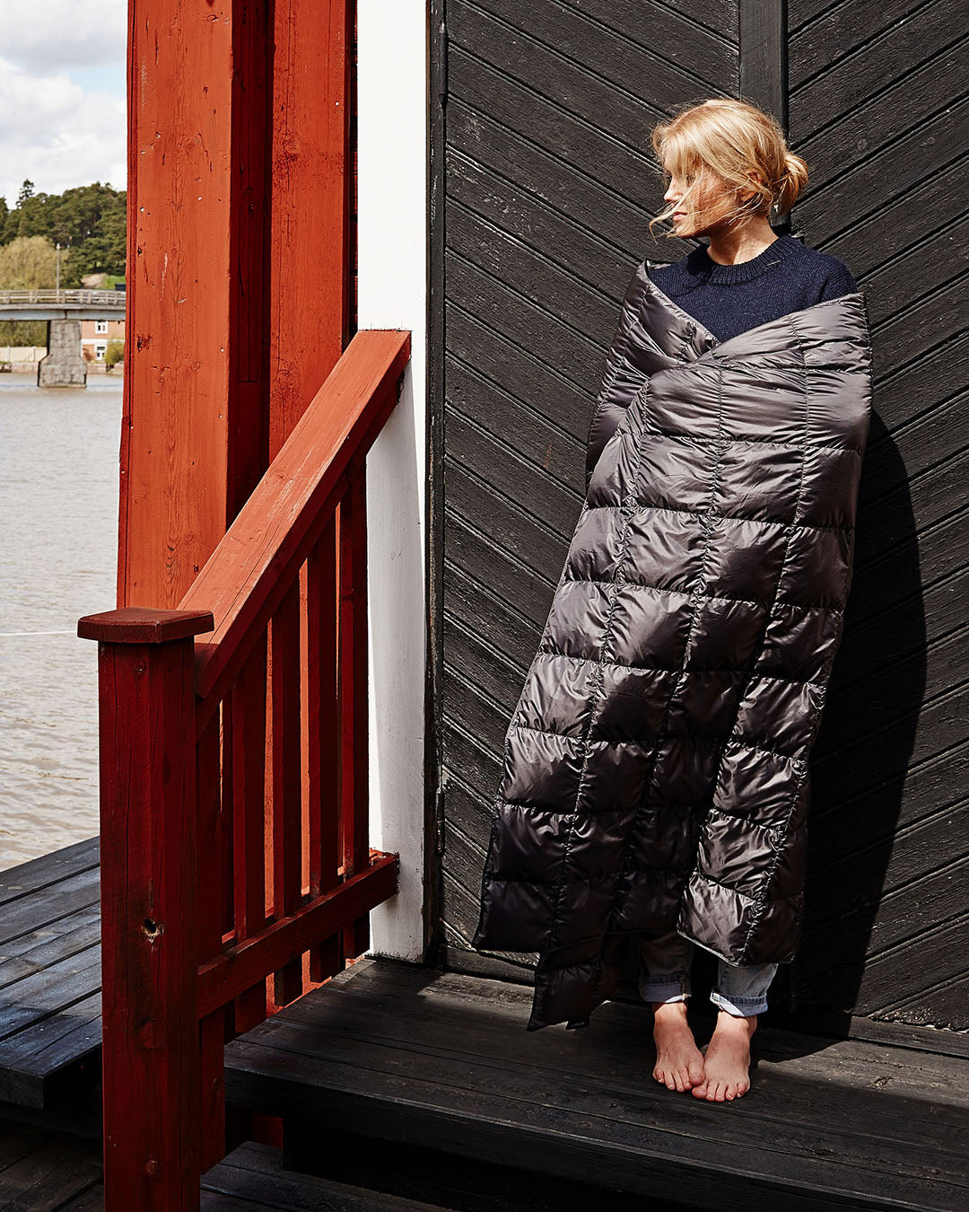 Henkilö seisoo paljain jaloin puuterassilla, kääriytyneenä tummanharmaaseen Kulkuri-untuvatorkkupeittoon (väri: antrasiitti). Hän nojaa mustaan lautaoveen ja katsoo vasemmalle. Taustalla näkyy jokimaisema ja vihreitä puita vastarannalla.